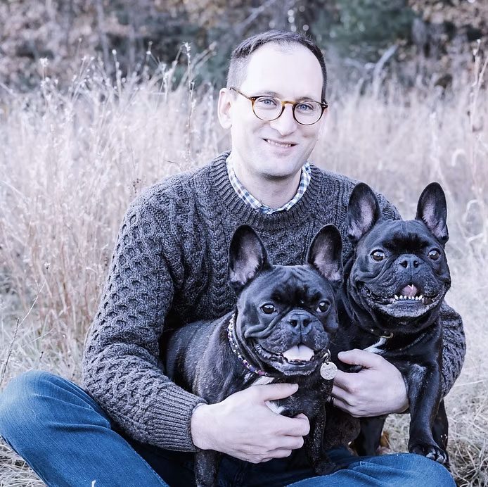 dr. christopher ryan sitting in the grass outside smiling and holding two black French Bulldogs